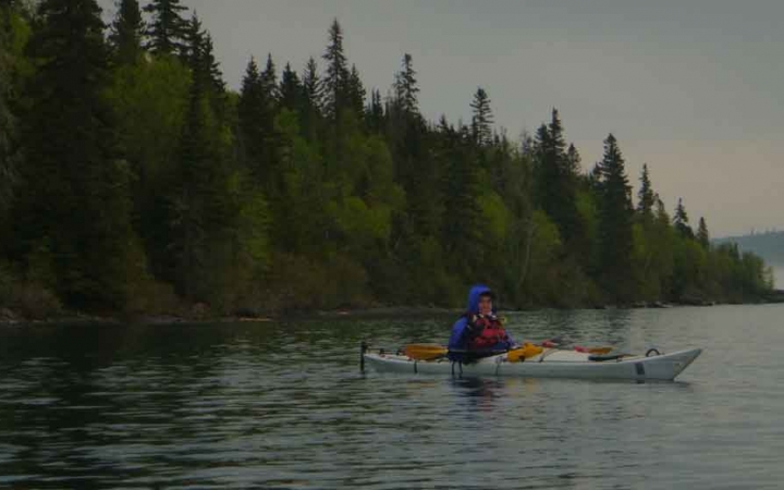 a student sits in a kayak on a calm lake lined with trees
